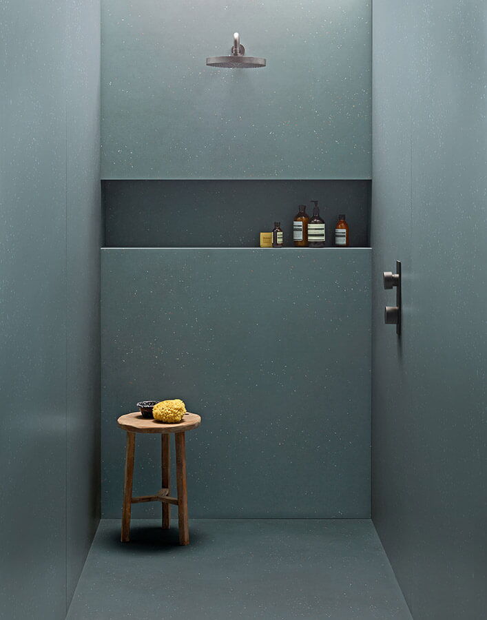begehbare Dusche mit grauen, großen Fliesen und Ablage mit Dusch-Utensilien, dazu ein Holzhocker