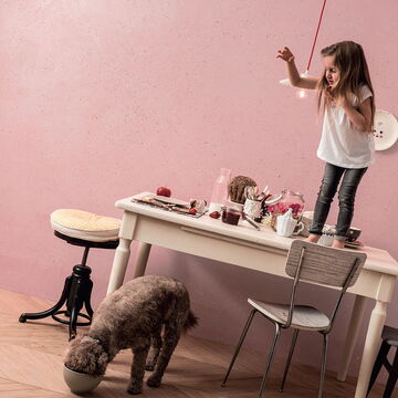 schräge Aufnahme eines kleinen Mädchens auf einem Tisch, davor ein Hund, der frisst; im Hintergrund rosa verputzte Wand