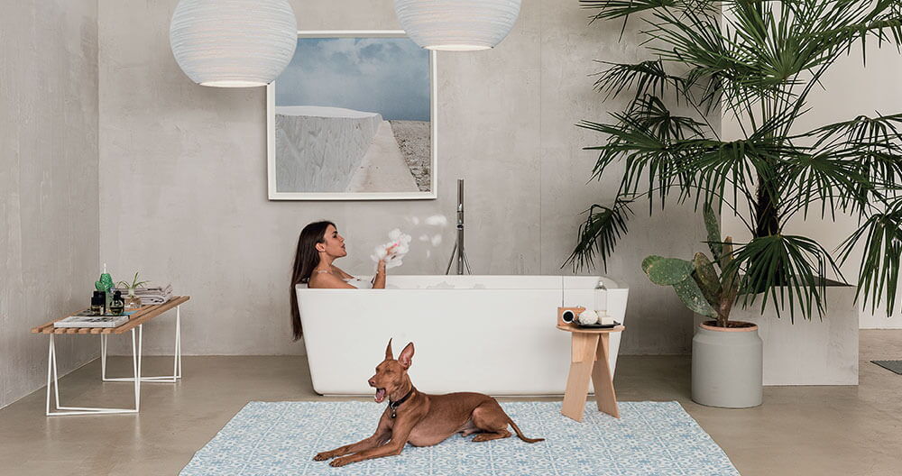 großer Raum mit freistehender Badewanne, in der eine Frau liegt, davor ein großer Hund auf einem Teppich, im Hintergrund eine grau verputzte Wand mit einem Bild