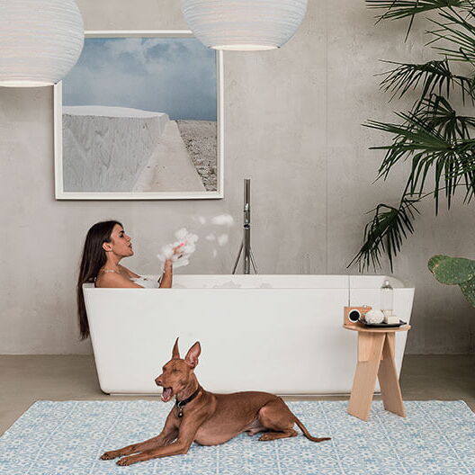 großer Raum mit freistehender Badewanne, in der eine Frau liegt, davor ein großer Hund auf einem Teppich, im Hintergrund eine grau verputzte Wand mit einem Bild
