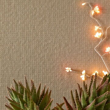 Wand mit sehr kleinen, quadratischen, weißen, unebenen Fliesen, davor Kakteen und eine Lichterkette