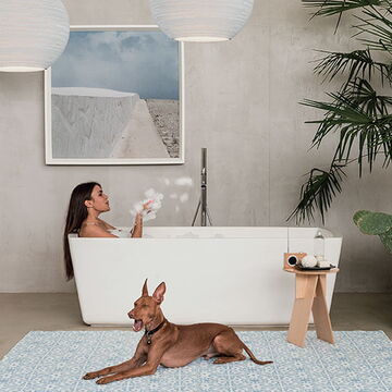 großer Raum mit freistehender Badewanne, in der eine Frau liegt, davor ein großer Hund auf einem Teppich, im Hintergrund eine grau verputzte Wand mit einem Bild