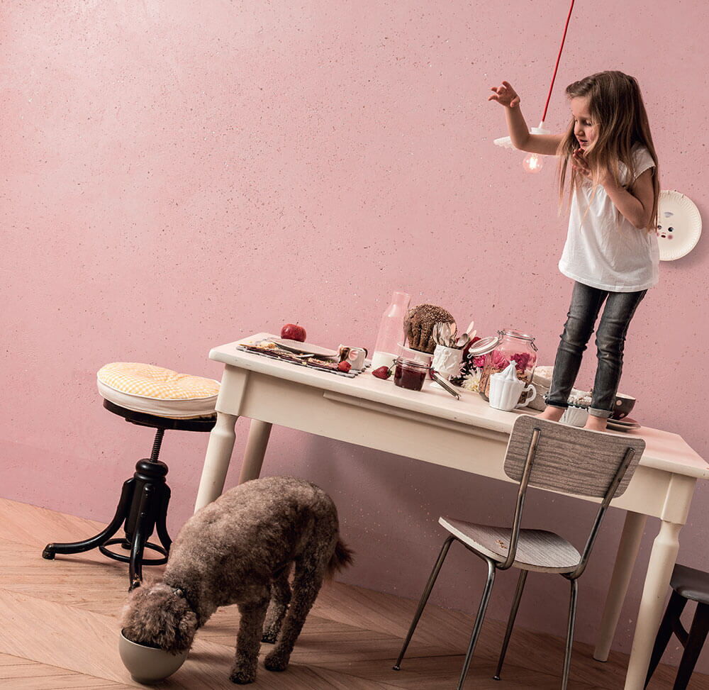 schräge Aufnahme eines kleinen Mädchens auf einem Tisch, davor ein Hund, der frisst; im Hintergrund rosa verputzte Wand